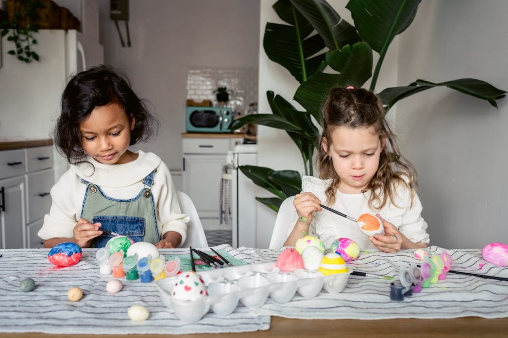 focused-multiracial-girls-painting-eggs-in-kitchen-7169309 Two children painting colorful Easter eggs at a kitchen table.